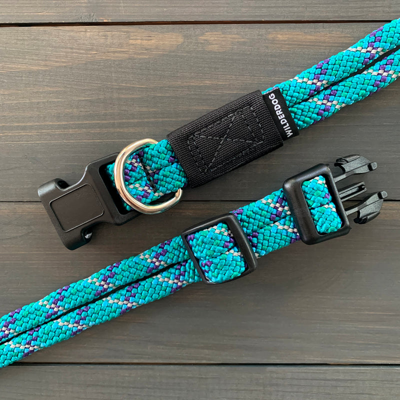 An Islander Reflective Collar, part of a blue and purple braided dog leash and collar set, is displayed on a wooden surface. The durable collar features a black plastic buckle, an adjustable strap, and heavy-duty hardware including a metal D-ring. A small tag with the brand name "Wilddog" is attached to the reflective leash near the clasp.